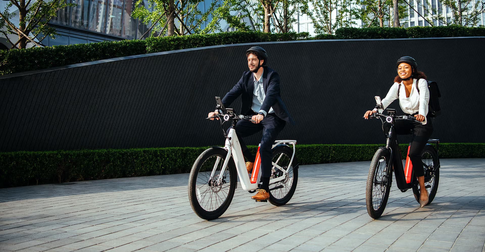 Two helmeted commuters riding NIU BQi-C3 Pro electric bikes through a landscaped city setting
