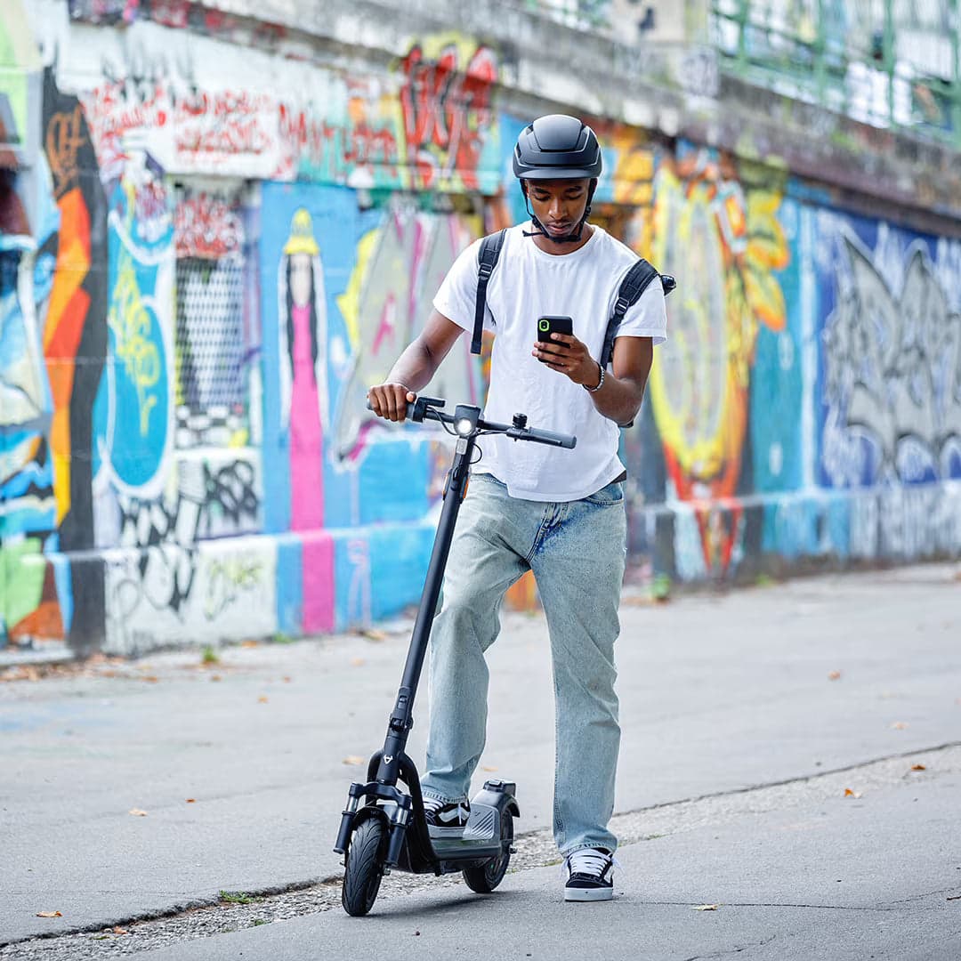 Helmeted rider standing with a NIU KQi 100 Series electric scooter beside a colorful city wall